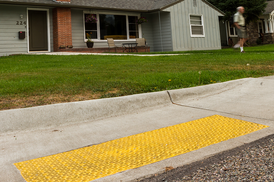 A zoomed in picture showing a detailed curb-ramp from a sidewalk to a street. In the background, a man continues walking away from a house.